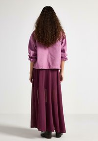 Kvinde med langt krøllet hår iført en lyserød jakke over en gennemsigtig, mørk lilla maxinederdel og sorte ankelstøvler, stående mod en hvid baggrund.