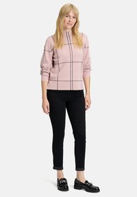 Helles rosa Sweater mit schwarzem Karomuster, rundem Ausschnitt und gerafften Ärmeln, kombiniert mit schwarzen hochgekrempelten Hosen und glänzenden schwarzen Schuhen.