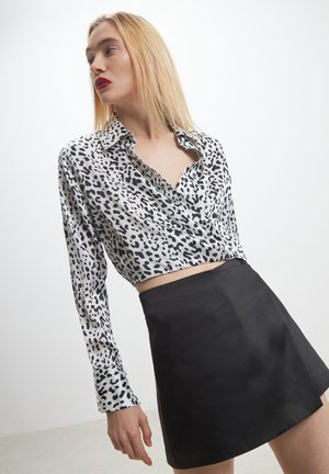 Une femme aux cheveux blonds porte un blouse en imprimé léopard noir et blanc ainsi qu'une jupe noire, regardant sur le côté devant un fond blanc uni.