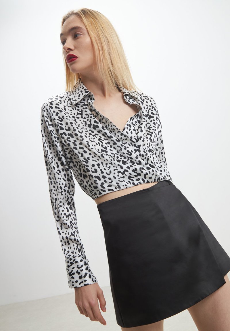 Donna con capelli biondi che indossa una blusa a stampa leopardata bianca e nera e una gonna nera, che guarda di lato su uno sfondo bianco semplice.