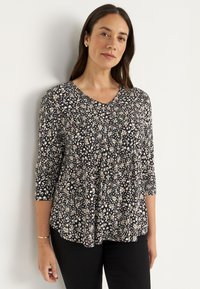 Zwarte top met 3/4 mouwen, v-hals, met een bloemenpatroon in wit en beige. De stof is zacht met een losse, vloeiende silhouette.