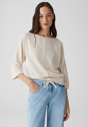 Maglione leggero beige con maniche a pipistrello, tessuto testurizzato, scollatura rotonda, abbinato a jeans azzurri con coulisse. Design semplice.