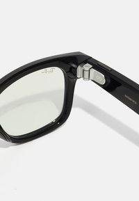 Gros plan sur la charnière des lunettes intelligentes Ray-Ban Meta noires avec un détail en métal argenté et des lentilles translucides sur un fond blanc.
