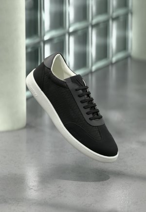 Pantof sport negru cu talpă albă, părți laterale texturate, șireturi negre și numele mărcii pe călcâi, prezentat pe un fundal modern din blocuri de sticlă.