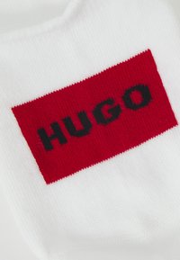 Fehér pamut zoknik, amelyek előtérbe helyezik a piros téglalap alakú foltot a "HUGO" fekete betűs felirattal. Puha tapintású, bordás kialakítású.