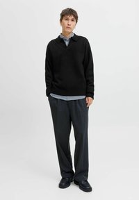 Sort strik sweater med V-hals, båret over en kravebluse med striber. Sammen med mørke, løstsiddende bukser og kraftige sorte sko.
