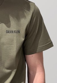 T-shirt in cotone verde oliva con scollo a giro e maniche corte, con "CALVIN KLEIN" stampato in nero sul lato sinistro del petto.