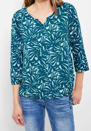 Vihreä pusero, jossa on vaaleansininen lehtikuvio, v-mallinen pääntie ja kolmannesosa hihat. Helmassa on sidottu yksityiskohta. Yhdistetty sinisten farkkujen kanssa.