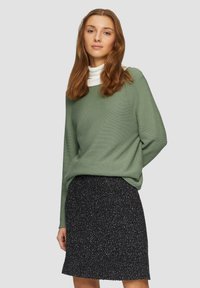 Hellgrüner gerippter Pullover mit Rundhalsausschnitt, über einem weißen Rollkragenpullover getragen, kombiniert mit einem schwarzen strukturierten Rock.