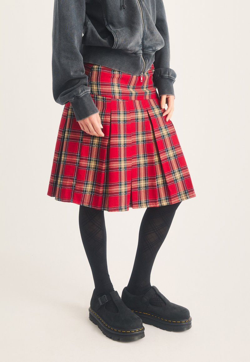 Person wearing a red plaid pleated skirt, black patterned tights, black buckle shoes, and a dark gray zip-up hoodie.