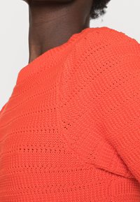 Maglione arancione lavorato a maglia con strisce testurizzate. Presenta un collo rotondo e cuciture visibili, che mettono in mostra un tessuto leggero e traspirante.