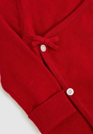 Sarkans adīts kardigāns ar sānslēga aizdari, ar baltām pogām un ribotām piedurknēm. Gluda tekstūra ar smalkām šuvēm.