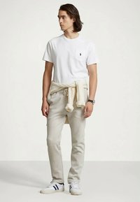 Młody mężczyzna ubrany w białą koszulkę, beżowe dresy, jasny sweter związany w talii oraz białe trampki w czarne paski, stoi i patrzy w lewo.