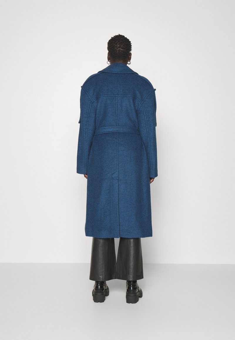 Długi niebieski wełniany płaszcz z paskiem w talii, dużym kołnierzem i opadającymi ramionami. Tył ma gładką teksturę i dopasowaną sylwetkę.
