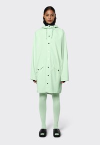 Lichtgroene regenjas van waterdicht materiaal, met een capuchon, twee voorzakken en zwarte drukknoopjes; gedragen over bijpassende leggings en zwarte sandalen.