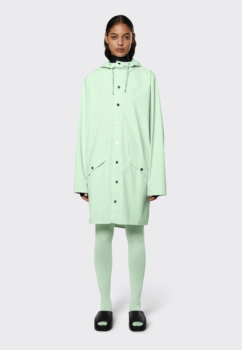 Lichtgroene regenjas van waterdicht materiaal, met een capuchon, twee voorzakken en zwarte drukknoopjes; gedragen over bijpassende leggings en zwarte sandalen.