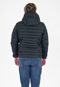 Man in een zwart gewatteerd jack met capuchon en blauwe jeans, staand met de rug naar de camera tegen een witte achtergrond.