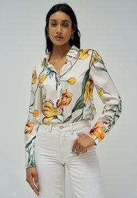 Camicia floreale realizzata in tessuto morbido, con fiori gialli e arancioni su uno sfondo crema, con chiusura frontale a bottoni e maniche lunghe.