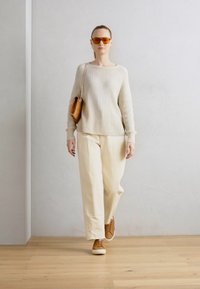 Pull en maille crème, pantalon large beige clair, baskets marron. Petit sac à main marron avec bandoulière en chaîne. Fond avec un mur de couleur claire.