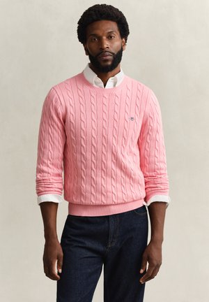 Man with beard wearing a white collared shirt under a pink cable-knit sweater and dark blue jeans, standing against a plain background.