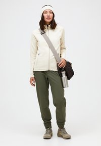 Helle beige Zip-Jacke, olivgrüne Hose, beige Schuhe, trägt eine graue Tasche und eine silberne Wasserflasche, mit einem weißen Stirnband.