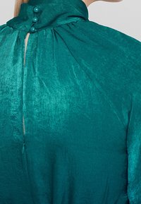 Tealblus med hög krage, featuring en knäppning bak och mjukt, glänsande tyg med plisserade detaljer.