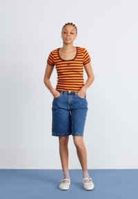 Oransje og brune stripete kortermet skjorte, blå denimshorts, grå sokker og lyse sko; avslappet passform med dyp halsing og sidelommer.