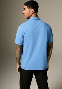 Světle modrá polo košile s klasickým límcem, krátkým rukávem a bočními rozparky, doplněná o černé kalhoty a s viditelnými tetováními na paži.