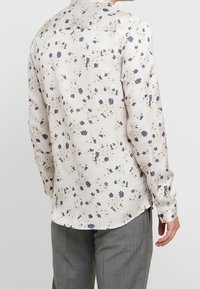 Light-colored long-sleeve shirt made of soft fabric, featuring a floral pattern in purple and beige. Back view with a collar and buttons.