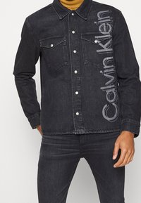 Chaqueta de denim negra con dos bolsillos en el pecho, botones a presión y gran texto en gris "Calvin Klein" en la parte frontal de forma vertical. Diseño ajustado.