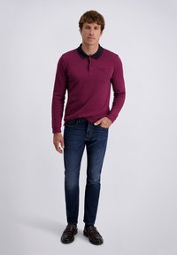 Bordeaux lange mouwen polo met een zwarte kraag, voorzien van een borstzak. Gecombineerd met donkerblauwe jeans en bruine instap schoenen met textuurdetail.