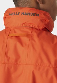 Cazadora naranja con cuello alto. Presenta un logo negro de "Helly Hansen", tejido texturizado y un pequeño bucle en la parte posterior para colgar.