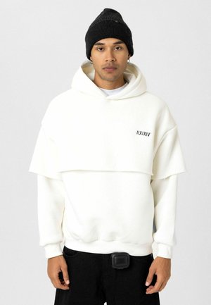 Witte oversized hoodie met gelaagd ontwerp en zwart logo, gecombineerd met zwarte broek. Model draagt een zwarte gebreide beanie en houdt een pouch vast.