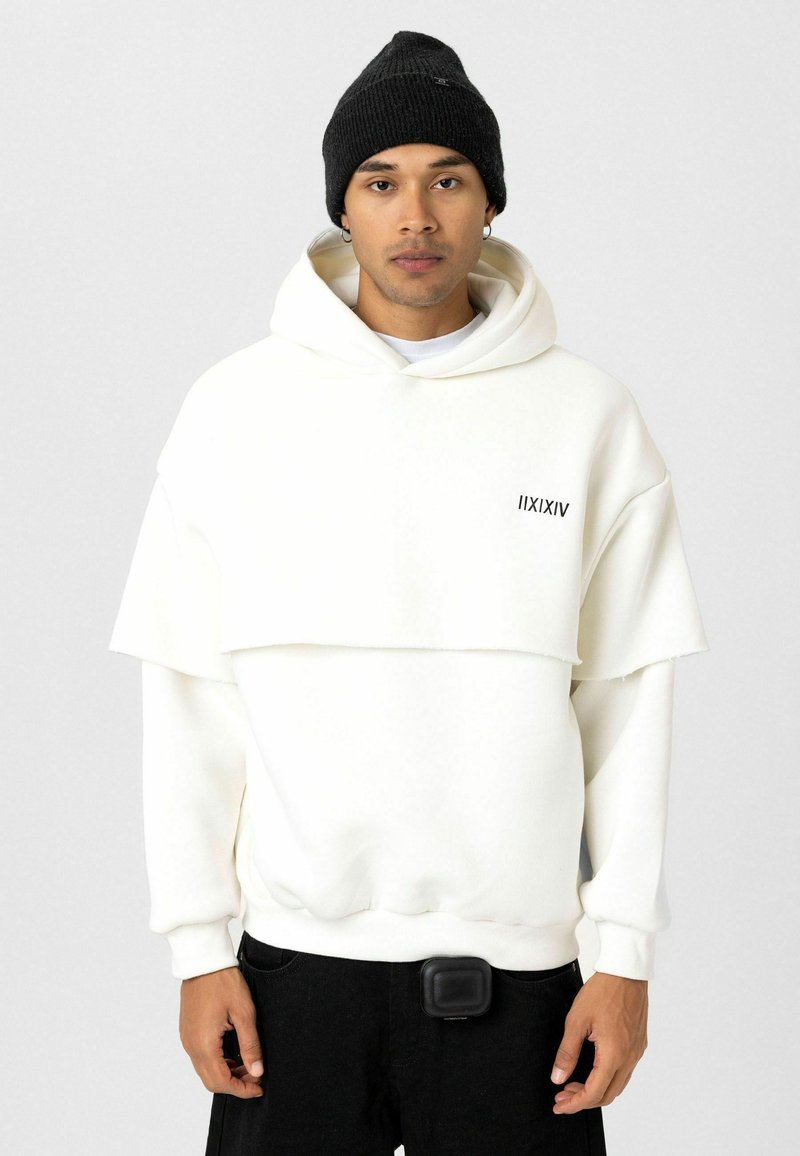 Witte oversized hoodie met gelaagd ontwerp en zwart logo, gecombineerd met zwarte broek. Model draagt een zwarte gebreide beanie en houdt een pouch vast.