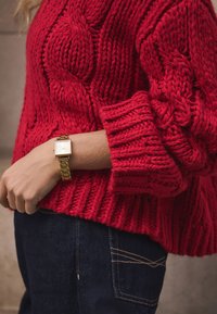 Red cable-knit sweater with ribbed cuffs, paired with a gold rectangular watch on a wrist and dark blue denim jeans.