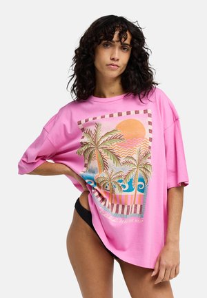 Femme aux cheveux bouclés et foncés portant un t-shirt rose oversize avec un motif palmier et coucher de soleil, associé à un bas de bikini noir.