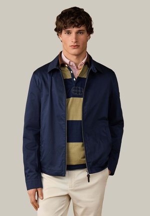 Young man wearing navy jacket over green and navy striped rugby shirt and beige pants standing against plain background.