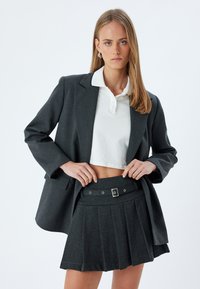 Grå skræddersyet blazer over en hvid kravecrop top, parret med en matchende grå plisseret nederdel med bælte og spænde detaljer.