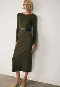 Abito midi fitted verde oliva con maniche lunghe, adornato da una cintura in pelle nera. La modella indossa scarpe con tacco alte di colore verde chiaro. Sfondo neutro.
