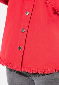 Chaqueta de mezclilla roja con botones a presión plateados y detalle de dobladillo deshilachado. Primer plano que muestra la textura y el contraste de color contra la tela gris.