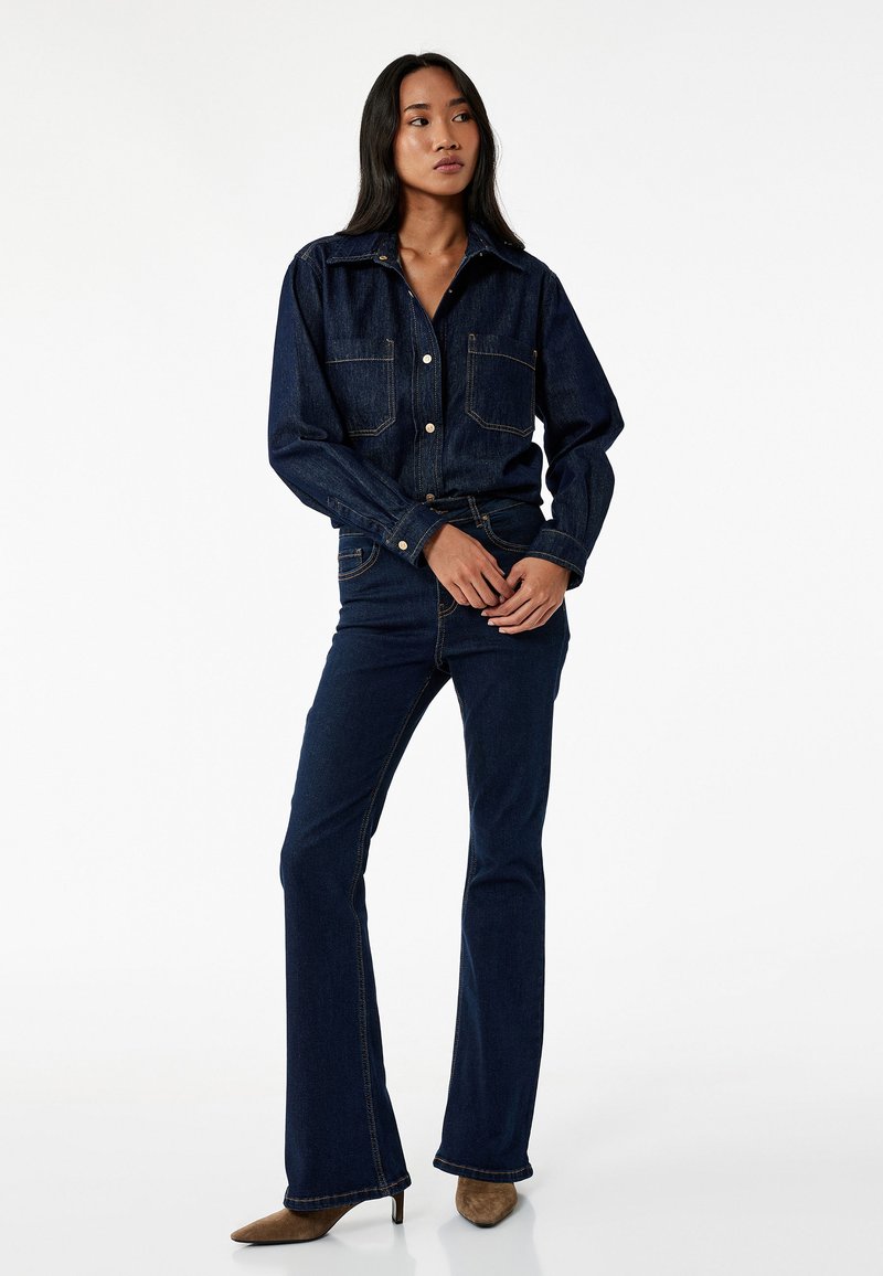 Mørkeblå denimjakke med to brystlommer, parret med flagrende mørkeblå denimjeans. Begge har kontrasterende syninger og en struktureret pasform.