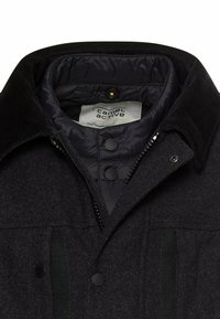 Schwarze Jacke mit einer strukturierten, wollähnlichen Außenseite, einem gesteppten Futter, Druckknöpfen und einem Reißverschluss vorne für Wärme.