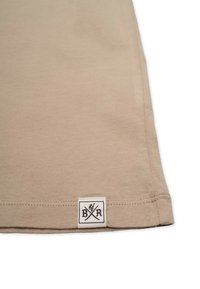 Beige cotton t-shirt featuring a rounded hem, side stitching, and a small branded label with black graphics on the lower edge.