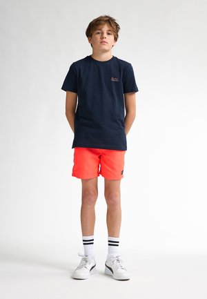 Marineblauwe katoenen T-shirt met ronde hals, gecombineerd met fel oranje shorts. Het model draagt witte sneakers en gestreepte witte sokken. Eenvoudig ontwerp.