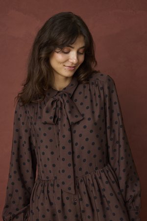Jeune femme aux longs cheveux foncés porte une robe marron à pois noirs et un nœud noué au col, regardant vers le bas avec un léger sourire.