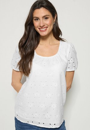 Kvinde med langt brunt hår smiler, iført en hvid kortærmet top med blomstrede hulmønstre og blå jeans mod en ensfarvet baggrund.