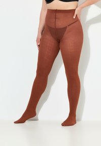 Leotardos marrones con textura, de cintura alta y patrón intrincado. La tela transparente se extiende hasta los pies, en contraste con una pretina sólida.