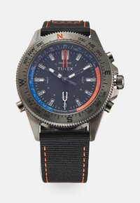 Montre Timex avec cadran analogique noir, boîtier en acier inoxydable argenté, bracelet en tissu noir, coutures orange et accents bleu-rouge sur la lunette.