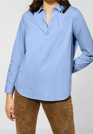 Mujer con una blusa celeste de manga larga con cuello y pantalones de pana marrones, de pie con una mano detrás de la espalda.