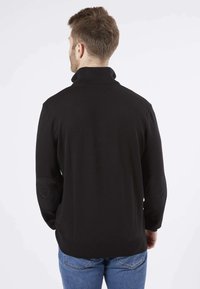 Pull en tricot noir à col montant, avec des patches aux coudes texturés et une coupe ajustée, porté avec un jean bleu.
